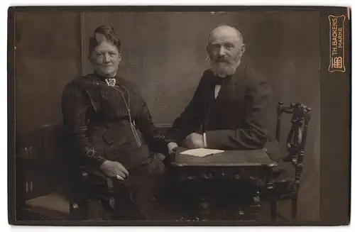 Fotografie Th. H. Backens, Marne, Älteres Paar in hübscher Kleidung am Tisch