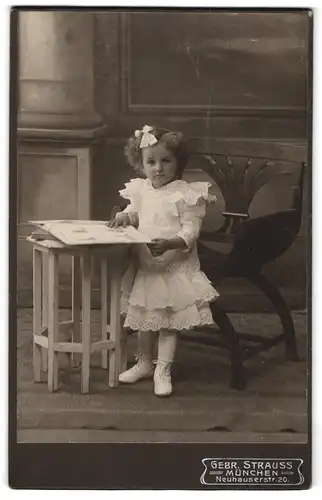 Fotografie Gebr. Strauss, München, Neuhauserstr. 20, Kind im weissen Spitzenkleid am Tisch mit Buch