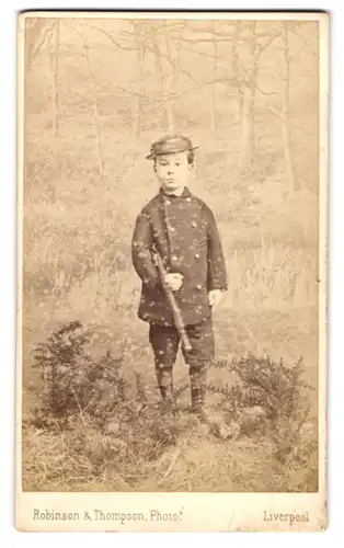 Fotografie Robinson & Thompson, Liverpool, Portrait junger Knabe als Jäger mit Gewehr vor einer Studiokulisse