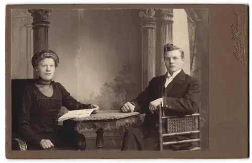 Fotografie Atelier Wretmark, Stockholm, Drottninggatan 68, Elegant gekleidetes Paar mit Buch am Tisch