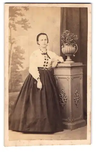 Fotografie H. Eckert, Prag, Junge Dame mit zurückgebundenem Haar im karierten Reifrockkleid