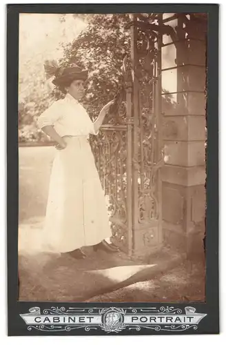 Fotografie unbekannter Fotograf und Ort, Dame in hellem Kleid mit Hut am Tor eines Parkeingangs