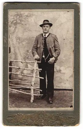 Fotografie Visite Portrait, Ort unbekannt, junger Mann in Tracht an einem Holzzaun stehend