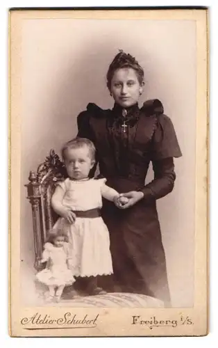 Fotografie Atelier Schubert, Freiberg i. Sa., junge Mutter im schwarzen Kleid mit Tochter samt Puppe, Perlenkette