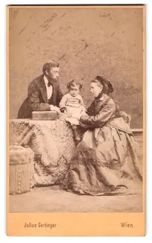 Fotografie Julius Gertinger, Wien, Mutter und Vater mit auf Tisch sitzendem Kleinkind, Mutterglück