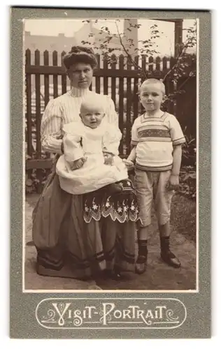 Fotografie unbekannter Fotograf und Ort, Mutter mit Kleinkind auf dem Schoss neben Sohn mit Spielzeug in der Hand