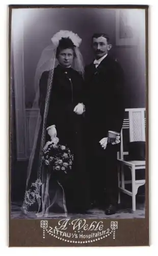 Fotografie A. Wehle, Zittau i. S., Hochzeitspaar im schwarzen Kleid und Anzug neben Stuhl mit Zylinder