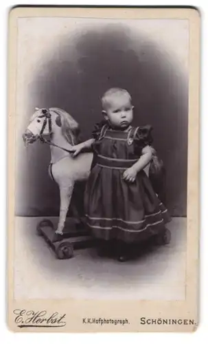 Fotografie E. Herbst, Schöningen, Kleinkind im Kleid steht vor dem Schaukelpferd auf Rollen