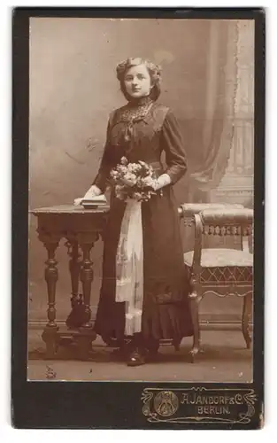 Fotografie A. Jandorf & Co., Berlin, Mädchen im Kleid mit Blumenstrauss neben Konsole zur Konfirmation