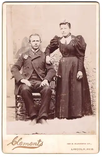 Fotografie Clements, Lincoln /Neb., Braut im schwarzen Kleid mit Puffärmeln neben ihrem Mann im Anzug