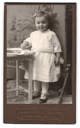 Fotografie Alfred Richter, Radebeul, süsses kleines Mädchen in entzückendem weissen Kleidchen an einem Tisch