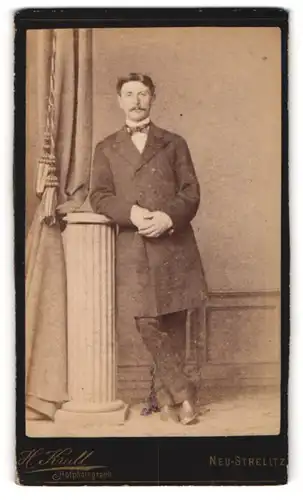 Fotografie H. Krull, Neu-Strelitz, Herr in zeitgenössischem Anzug mit Schnauzbart an einer Säule