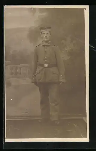 Foto-AK Soldat in Uniform mit Krätzchen und Brille vor Studiokulisse