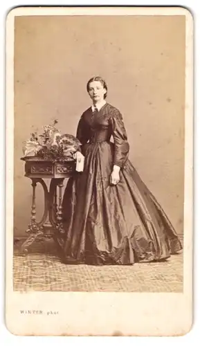 Fotografie M. L. Winter, Prag, Portrait junge Dame im Reifrockkleid mit Blumenbuket auf dem Tisch