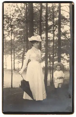Fotografie unbekannter Fotograf und Ort, junge Mutter im weissen Kleid mit Hut und Schirm samt Kind im Wald