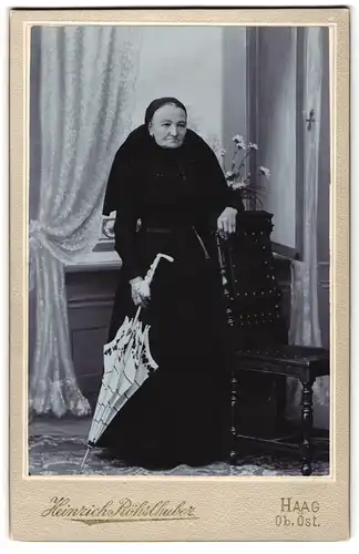 Fotografie Heinrich Röhslhuber, Haag / Ob. Öst., ältere Dame im schwarzen Kleid mit Kopftuch und Schirm