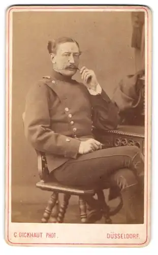 Fotografie G. Dickhaut, Düsseldorf, Soldat in Uniform posiert sitzend im Atelier und zwirbelt seinen Bart