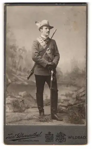 Fotografie Karl Blumenthal, Wildbad, Portrait Jäger mit geschultertem Gewehr und Munitionsgurt, Säbel und Federhut