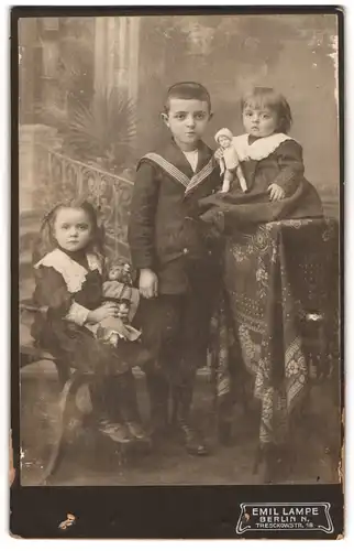Fotografie Emil Lampe, Berlin, drei junge Kinder im Matrosenanzug und in Kleidern mit ihren Puppen im Atelier