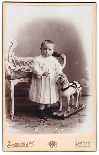 Fotografie Samson & Co., Stuttgart, Kleinkind im weissen Kleidchen mit Spielzeug Pferd im Atelier