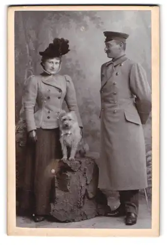 Fotografie Franz Tausch, Amberg, Portrait Soldat in Uniformmantel nebst Frau und ihrem Hund auf einem Stein