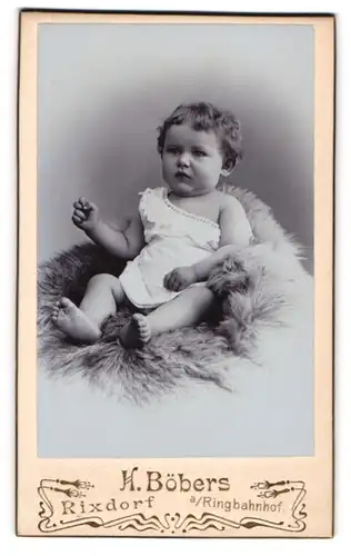 Fotografie H. Böbers, Berlin-Rixdorf, Drolliges Kleinkind im weissen Kleidchen auf Pelz sitzend