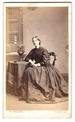 Fotografie F. Spalding, Chelmsford, Portrait junge englische Dame im Reifrockkleid posiert sitzend am Tisch