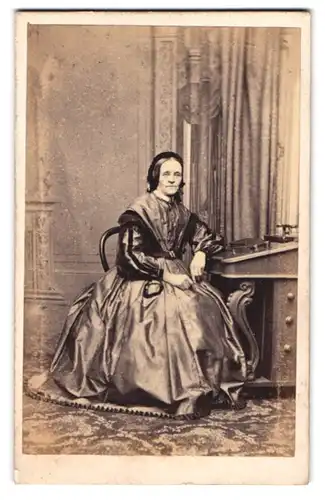 Fotografie E. Gregson, Halifax, ältere Engländerin im seidenen Kleid mit Haube sitzend am Sekretär