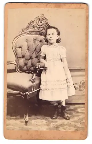 Fotografie M. S. Simson, Dillingen, kleines Mädchen im weissen Kleid mit Locken stehend neben einem Sessel