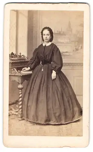 Fotografie F. Halm, Constanz, Portrait junge Frau im schwarzen Reifrockkleid stehend am Sekretär