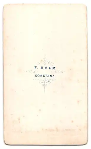 Fotografie F. Halm, Constanz, Portrait junge Frau im schwarzen Reifrockkleid stehend am Sekretär
