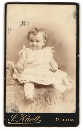 Fotografie S. Knott, Oldham, süsses kleines Mädchen im weissen Kleidchen auf Fell sitzend