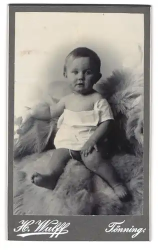 Fotografie H. Wulff, Tönning, Portrait süsses Baby im weissen Hemdchen auf Fell sitzend