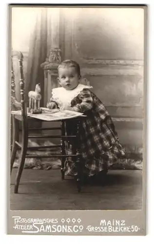Fotografie Samson & Co., Mainz, Grosse Bleiche 50, Kleinkind im karierten Kleid mit Spielzeugpferd
