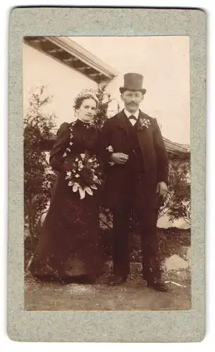 Fotografie unbekannter Fotograf und Ort, Eheleute im schwarzen Brautkleid und im Anzug mit Zylinder, Ansteckblume