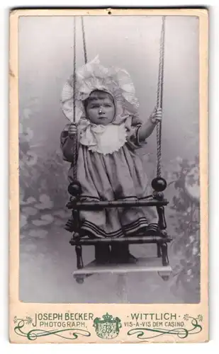 Fotografie Joseph Becker, Wittlich, Weinerliches Kleinkind mit weissem Häubchen in einer Schaukel stehend