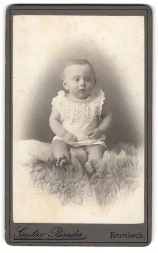 Fotografie Gustav Baader, Krumbach, Niedliches Baby im weissen Kleid auf einer Schafswolle