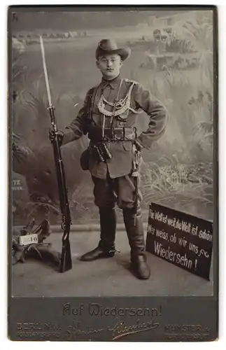 Fotografie Portrait Atelier Schubert, Berlin, junger DSWA-Soldat in Uniform mit Südwester, Schützenschnur und Fernglas