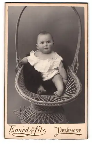 Fotografie Enard & fils, Delemont, niedliches Kleinkind im weissen Leibchen sitzt in einem Weidenkorb
