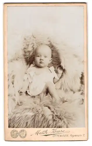 Fotografie Matth. Kruse, Ottensen, Papenstr. 7, Portrait süsses Baby im weissen Hemdchen auf Fell sitzend