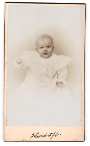 Fotografie J. Enard e fils, Delemont, Baby mit erhobenem Arm im Taufkleid