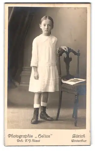 Fotografie Atelier Helios, Zürich, Bahnhofstr. 110, Portrait hübsches Mädchen im Kleid am Stuhl stehend