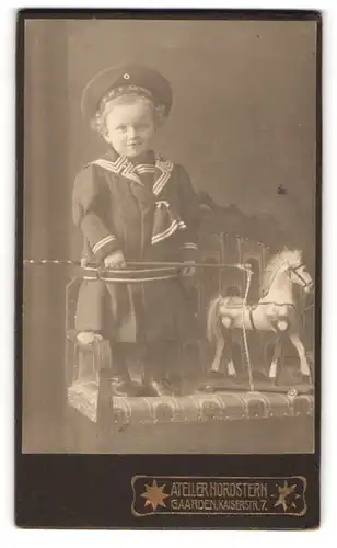 Fotografie Atelier Nordstern, Gaarden, niedliches Mädchen im Kleid mit Mützenband SMS Arcona, Spielzeugpferd