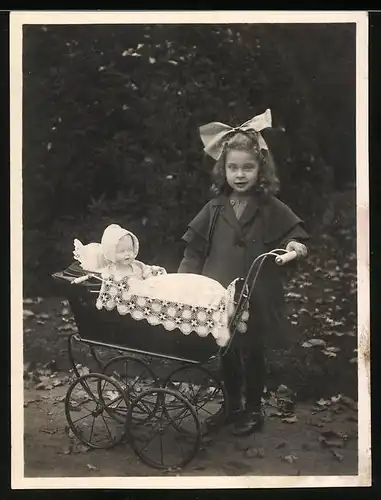 Fotografie Mädchen mit Haarschleife nebst Puppe im Puppenwagen