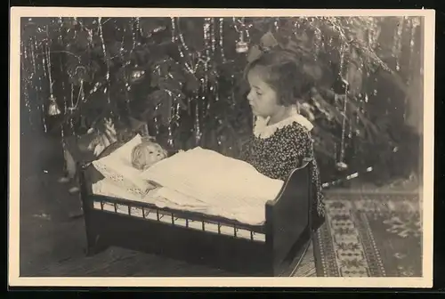 Fotografie Weihnachten, Mädchen mit Puppe im Puppenbettchen neben Weihnachtsbaum