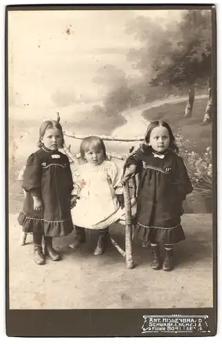 Fotografie Ant. Hillenbrand, Schwabmünchen, drei kleine Mädchen in Kleidern auf einer Holzbank vor einer Studiokulisse