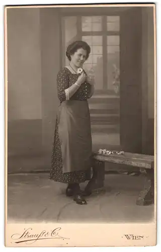 Fotografie Strauss & Co., Wien, Portrait Frau Hella Müller im Blümchenkleid mit Schürze vor einer Studiokulisse, 1914