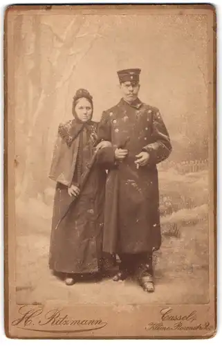 Fotografie H. Ritzmann, Cassel, Eisenbahner in Uniform mit Mantel und Mütze nebst Frau in einer Winter Studiokulisse