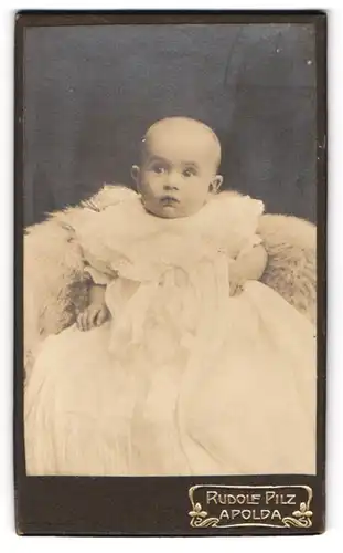 Fotografie Rudolf Pilz, Apolda, Ackerwandstrasse 34, Kleinkind im weissen Kleid sitzt auf Fell