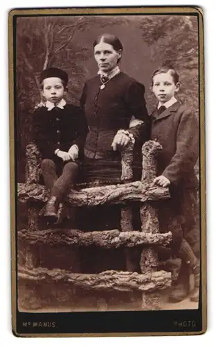 Fotografie J. M. Manus, Edinburgh, 13 High Street, Portrait stolzer Mutter mit zwei Buben am Holzzaun stehend